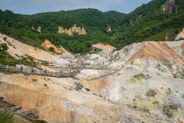 Tourists walking on wooden path through volcanic landscape in green mountain area at Noboribetsu Jigokudani Valley, Noboribetsu, Hokkaido, Japan