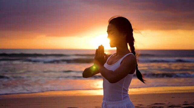 A silhouetted woman practicing yoga and meditation on the beach at sunset. Person praying with hands together by the ocean. Wellness and mindfulness concept
