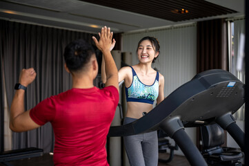 Fototapeta premium Fitness trainer man clapping hands high five with an asian woman client running on treadmill in gym