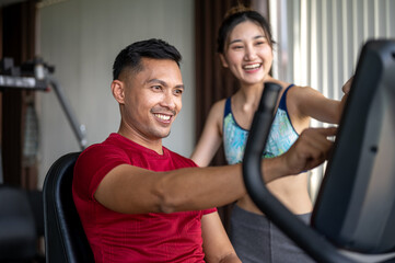 Fototapeta premium Smiling fitness trainer man teaching pretty asian woman client how to use exercise equipment in gym