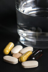 A glass of water beside assorted vitamin and supplement capsules on a dark background, symbolizing health, wellness, and daily nutrition.
