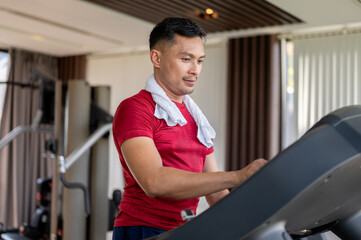 Asian middle aged healthy man with a towel adjust speed while running exercising on treadmill in gym