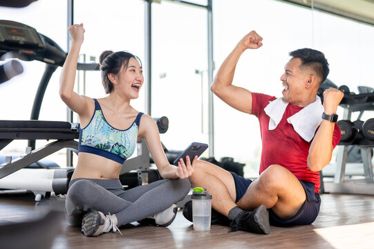 Asian woman client and fitness coach trainer a raising fist sitting on floor after exercising in gym