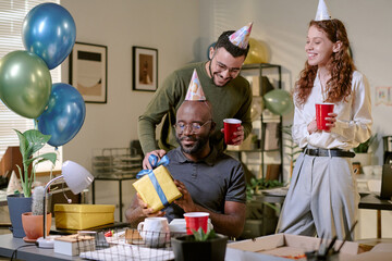 Black middle aged man sitting at desk opening gift box, while young adult Caucasian man and young adult Caucasian woman standing behind him smiling and holding party cups during office celebration