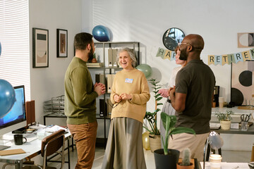 Senior Caucasian woman smiling and talking with middle aged Caucasian man and Black man during retirement celebration in office setting, people standing and interacting near decorations