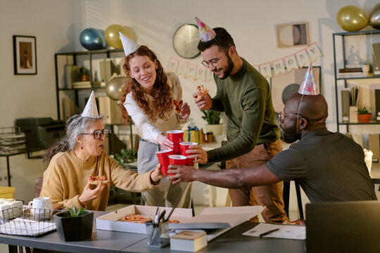 Group of diverse young adult and middle aged men and women wearing party hats celebrating birthday in office, holding pizza and raising red cups in toast, smiling and interacting together