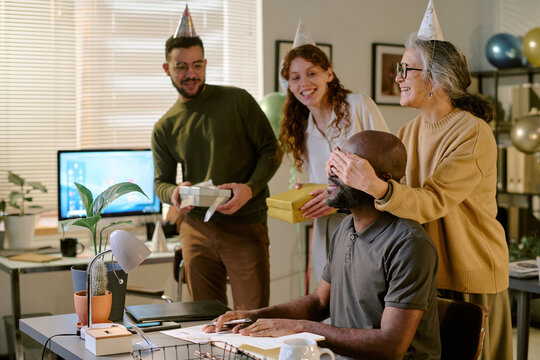 Middle aged Black man sitting at desk being surprised by middle aged Caucasian woman covering his eyes, while young adult multiethnic colleagues standing nearby holding gifts wearing party hats