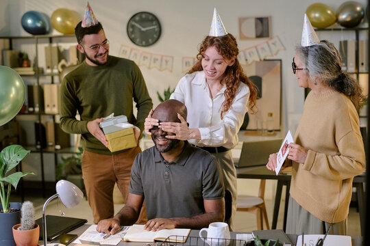Middle aged Black man sitting at desk being surprised by young Caucasian woman covering his eyes, while young man and senior woman standing nearby holding gifts during office celebration