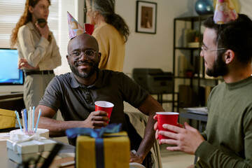 Black middle aged man wearing party hat sitting in wheelchair holding red cup, smiling at young Caucasian man during indoor birthday celebration with gifts and cake on table