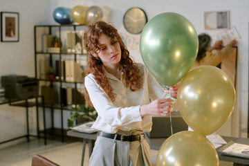 Young adult Caucasian woman arranging green and gold balloons in modern office setting, focusing on decoration task while another in background preparing wall banner for celebration
