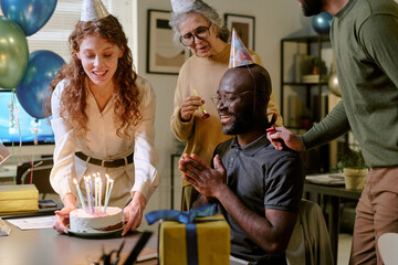 Middle aged Black man sitting at table smiling while young Caucasian woman presenting birthday cake with candles, senior Caucasian woman and young Caucasian man celebrating together
