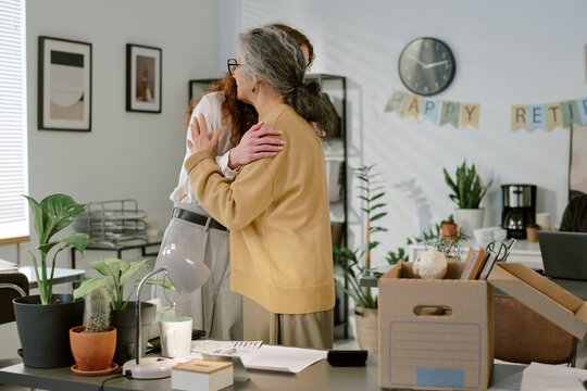 Senior Caucasian woman embracing young adult Caucasian woman in office setting, celebrating retirement with decorations and packed box on desk, both standing near workspace