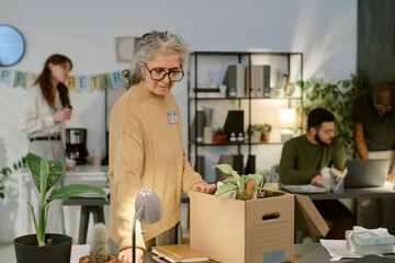 Senior Caucasian woman packing personal belongings into cardboard box at office desk, colleagues in background interacting near retirement banner, indoor workplace setting