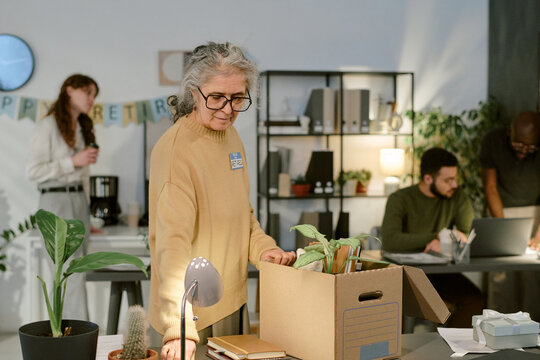 Senior Caucasian woman packing personal belongings into cardboard box at office desk, colleagues in background interacting near retirement banner, indoor workplace setting