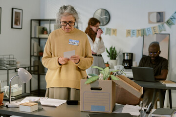 Senior Caucasian woman wearing glasses standing at desk reading card during office retirement celebration, cardboard box with personal belongings on table, colleagues working in background