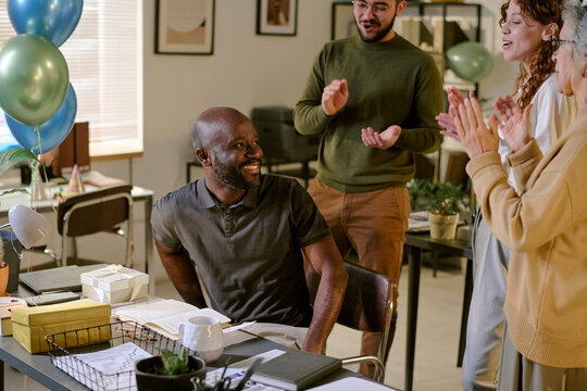 Middle aged Black man sitting at desk smiling, while diverse group of young adults and senior Caucasian woman standing nearby clapping and celebrating in office setting with balloons and gifts - Powered by Adobe