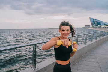 A woman intensively trains with dumbbells against the backdrop of a vast ocean, embodying strength...