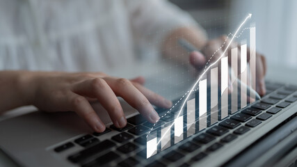 A close-up of a person typing on a laptop while a digital graph displays upward trends,...
