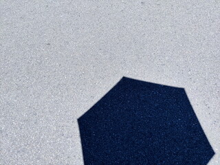 ザラザラとしたアスファルトのグレーの地面の上に濃いブルーの六角形のような形をした影が落ちている抽象的な構成