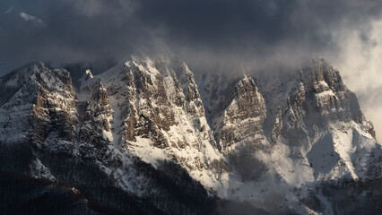 Aerial view of jagged, snow-dusted mountain peaks pierce through swirling clouds, their rocky faces kissed by the sun's golden light, Minucciano, Tuscany, Italy.