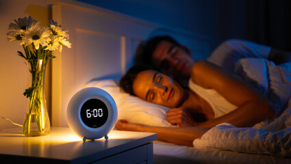 Couple wake up with the help of a smart alarm clock with sunrise simulation in a cozy bedroom. Modern wake up light technology for healthy sleep, natural morning routine, and stress-free lifestyle.