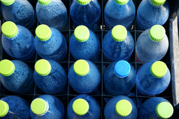 A neatly arranged set of blue plastic bottles with green caps, covered in condensation droplets, sits in a crate. The cool tones and water beads emphasize freshness and hydration, evoking the feeling 