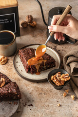 Chocolate spongy brownie cake with walnuts with hand drizzling caramel glaze by spoon and coffee on brown table background. Two pieces of chocolate brownie cake