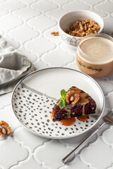 Chocolate spongy brownie cake served with walnuts and mint leaves on top and coffee on white tile background. A piece of chocolate brownie cake
