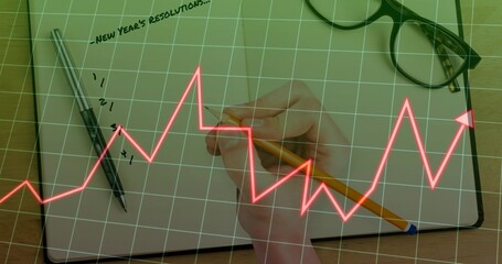 Hand writing New Year's resolutions on notebook at desk, with pencil, pen under grid graph overlays
