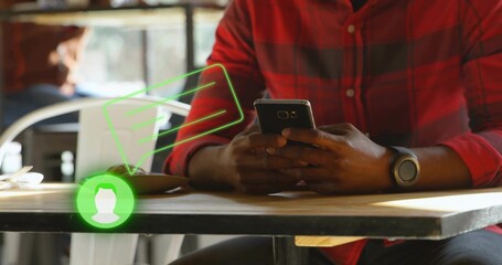 Man wearing plaid shirt checking smartphone, typing on laptop at wooden cafe table, with chat icons