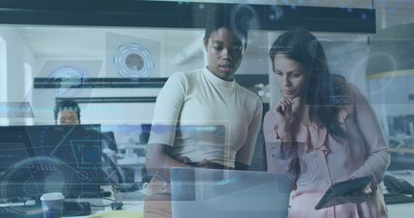 Collaborating women in work attire examining laptop in office with monitors, tablet and UI overlays