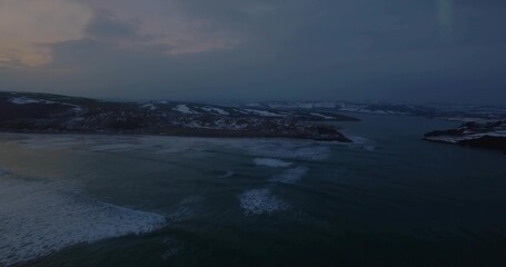 Rolling ocean waves gently lapping rocky snow-dusted shoreline at twilight, pastel sky