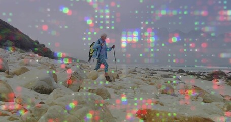Hiking senior woman in blue jacket navigating rocky coast beside sea, trekking poles and backpack