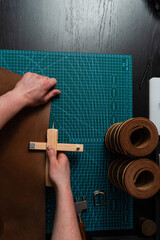Craftsman preparing leather strap using wooden cutter on mat. Handmade leather belt preparation with wooden strap cutter on cutting mat. Artisan is cutting leather strip with wooden tool.