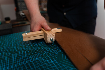 Craftsman preparing leather strap using wooden cutter on mat. Handmade leather belt preparation with wooden strap cutter on cutting mat. Artisan is cutting leather strip with wooden tool.