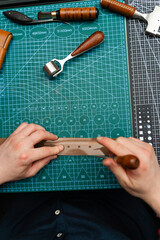 Crafting belt holes with punch tool on leather strap. Punching and detailing leather belt holes. Artisan punching holes into a leather strap for a handmade belt on a green cutting mat.