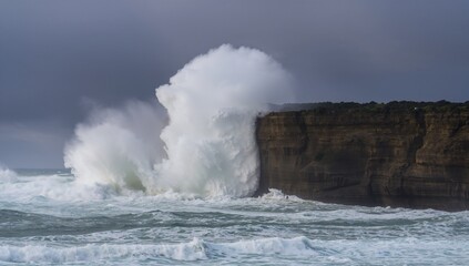 Obraz premium Crashing wave hitting rocky cliff at coast under gray sky, with foam-topped sea and white spray