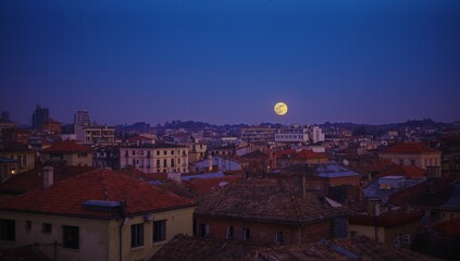 Rising full moon casting gentle glow over skyline at dusk, with tiled roofs, antennas and chimneys