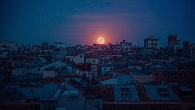 Rising full moon casting orange halo over multi-story rooftops at night, with chimneys and antennas - Powered by Adobe