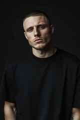 Fototapeta premium Intense portrait of a man with a short haircut and stubble wearing a black shirt against a dark background.