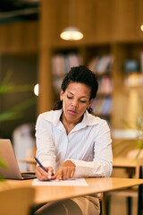 Obraz premium African-American Woman Focused While Writing at Desk with Laptop in Modern Office