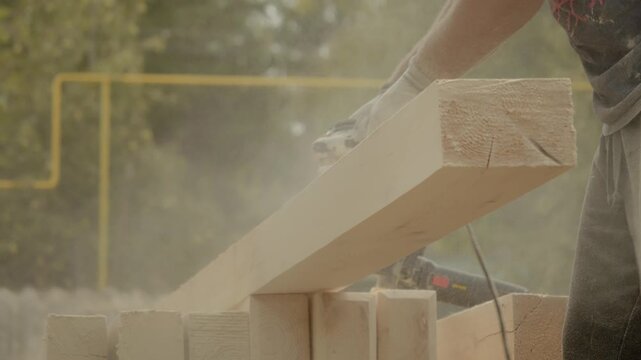 A skilled worker is sanding wooden beams with an angle grinder, creating a smooth finish in a sunny outdoor area. Dust fills the air as the tool rapidly shapes the wood.