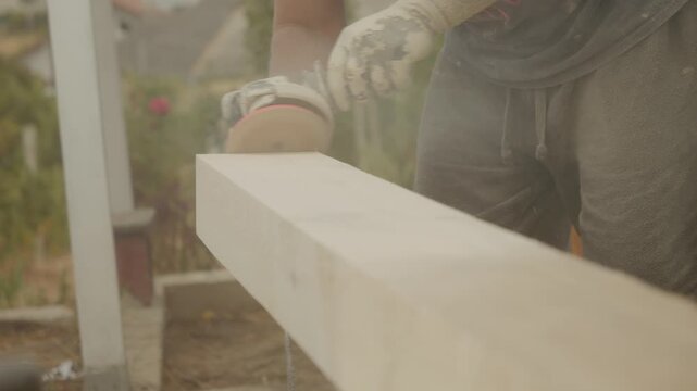 A worker is sanding wooden beams using an angle grinder in an outdoor workshop. Dust fills the air as the worker smooths the surface of the wood beams.