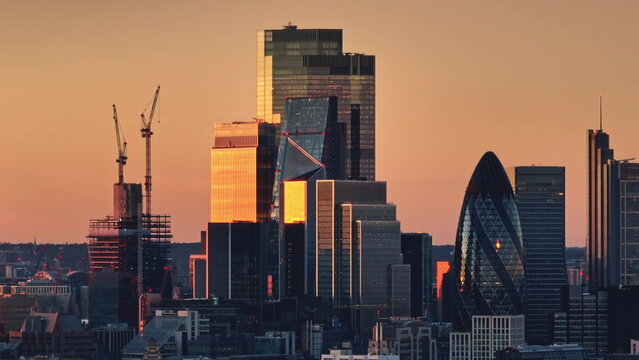 Bright orange sunset light over modern glass skyscrapers in London's financial district, iconic buildings like the Gherkin and active construction cranes. Business center skyline. Drone flight