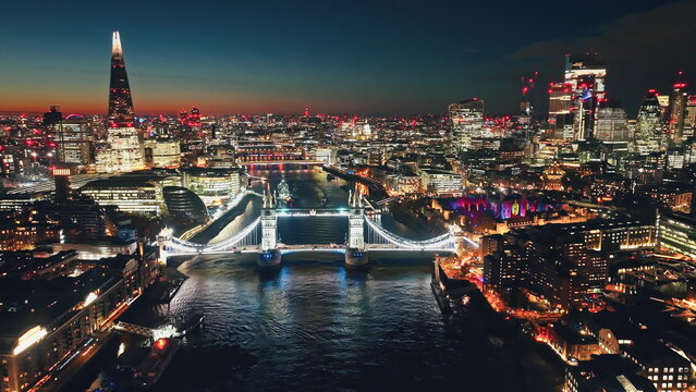 UK, London: Aerial view of famous Tower Bridge at night, The Shard and other landmarks along the Thames River, creating a stunning urban cityscape in colorful evening lights. Drone flight panorama