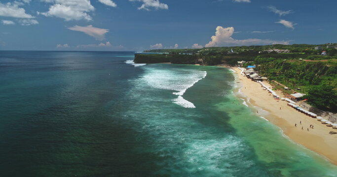 Stunning aerial view of Dreamland Beach in Bali, Indonesia, featuring vibrant turquoise waters, gentle waves on white sand, and lush green cliffs under a bright blue sky