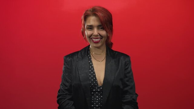 Woman smiling with visible cleavage and layered necklaces wearing polka dot dress and black blazer in red studio; confidence poise.
