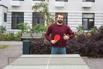Man in a burgundy sweatshirt and jeans standing by an outdoor table tennis table, holding a red...