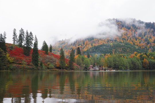 A view across a calm lake of a lakeside building and hills covered in colorful red and orange autumn foliage under a cloudy sky