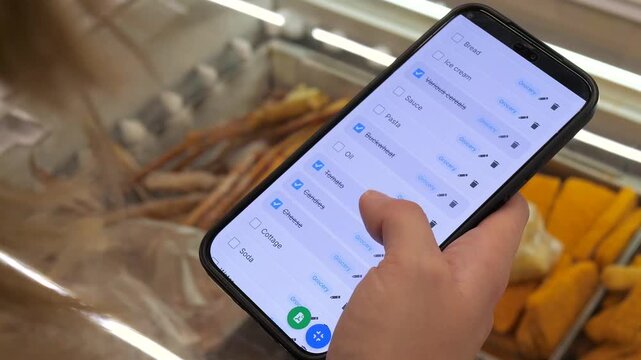 A shoppers hands in a supermarket navigate a digital grocery list on a smartphone, with some items already crossed off while others remain pending. 